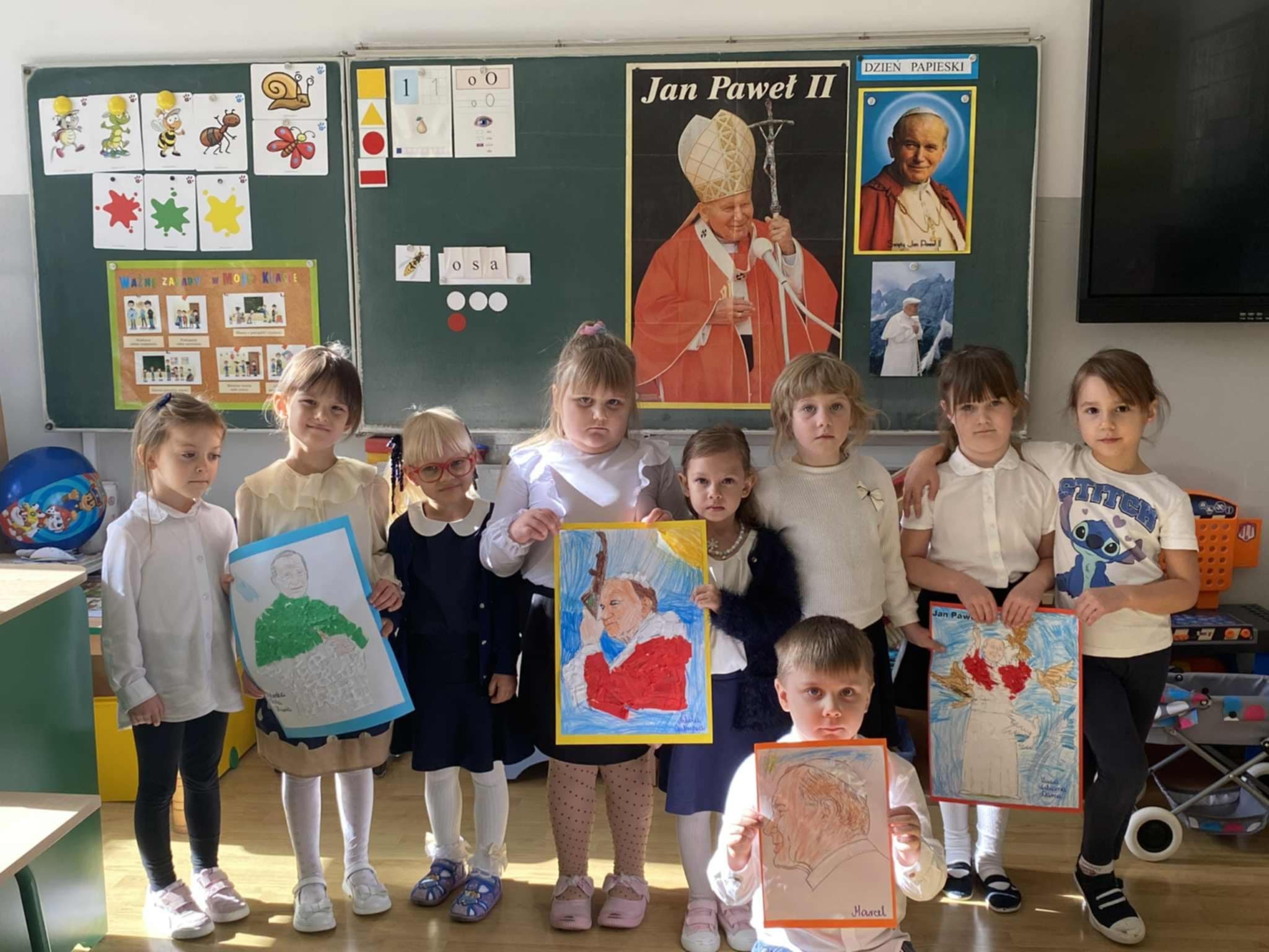 Dzieci trzymają w rękach wykonane przez siebie prace plastyczne przedstawiające Jana Pawła II.